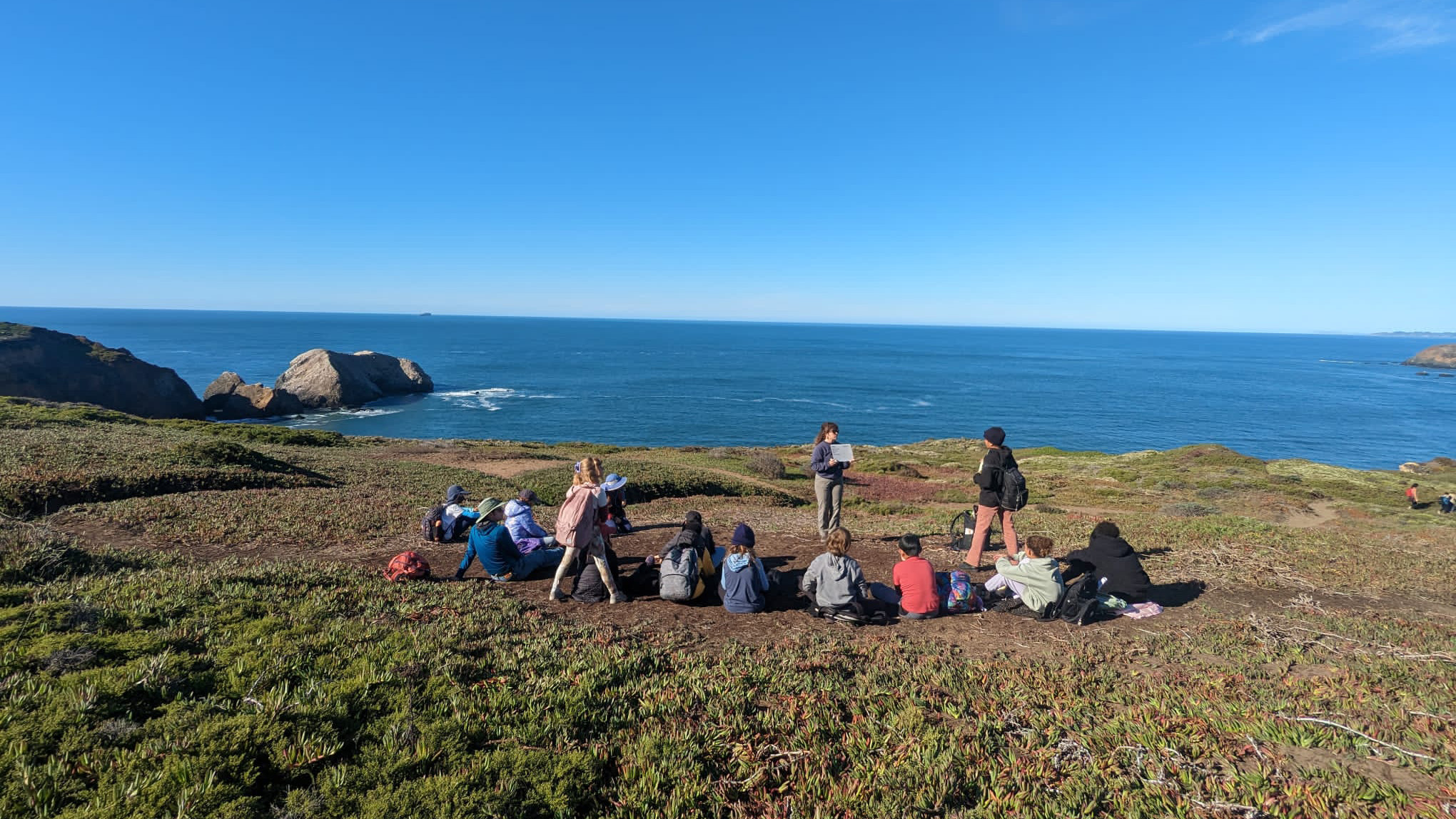 Group of students with their NatureBridge educator gathered in a circle near an ocean cliffside