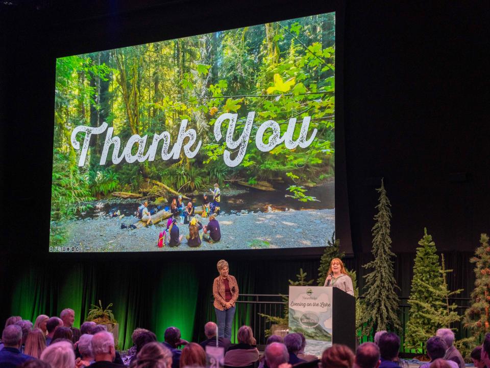 Two adults standing on a stage in front of an audience with a large projector screen reading "Thank you" above them