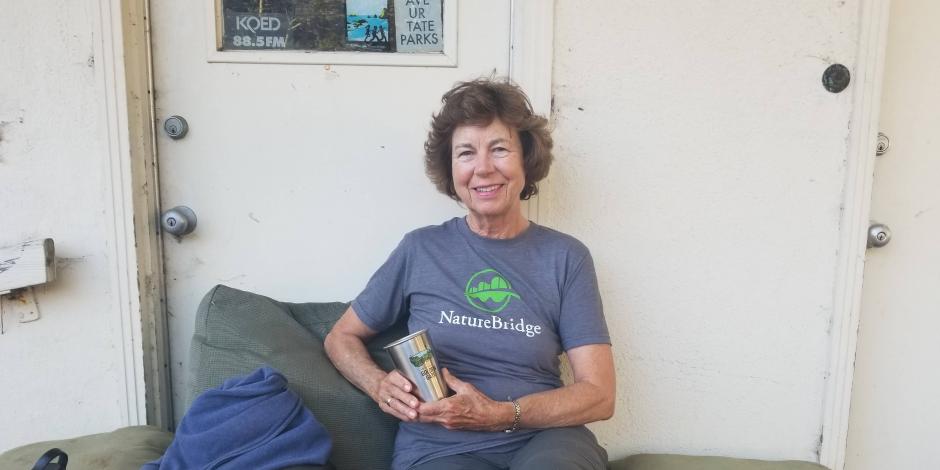 NatureBridge educator Ingrid Apter sitting against a white wall with a cup in hand sitting on green cushions