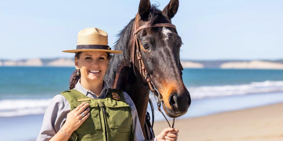 Julie, dressed in a green ranger's vest and a yellow broad-brimmed hat, stands on a beach in front of the ocean while holding the reigns of a dark brown horse.