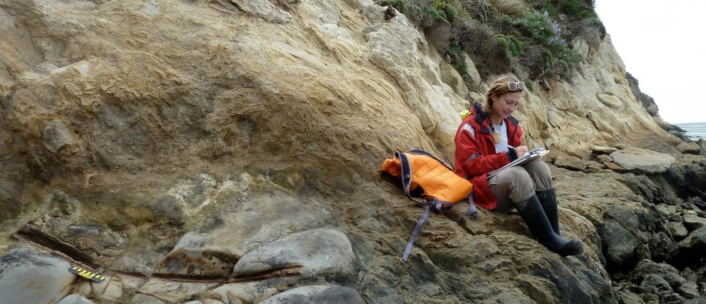 Lilly sits on a rocky shoreline wearing a rain jacket and tall rubber boats as she makes notes on a clipboard